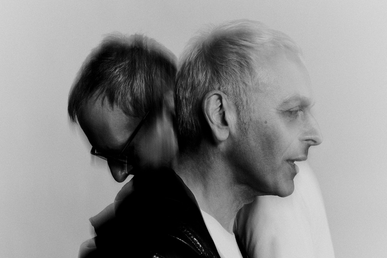 Black and white portrait of electronic music duo Underworld leaning back to back in side profile with soft motion blur.